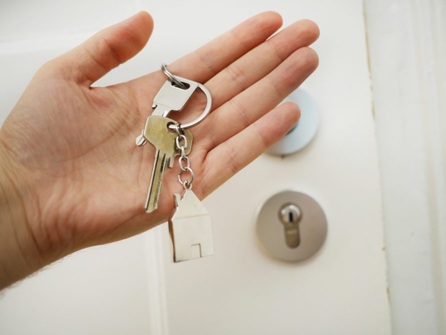 dwa klucze z breloczkiem trzymane w otwartej dłoni na tle drzwi do domu modułowego